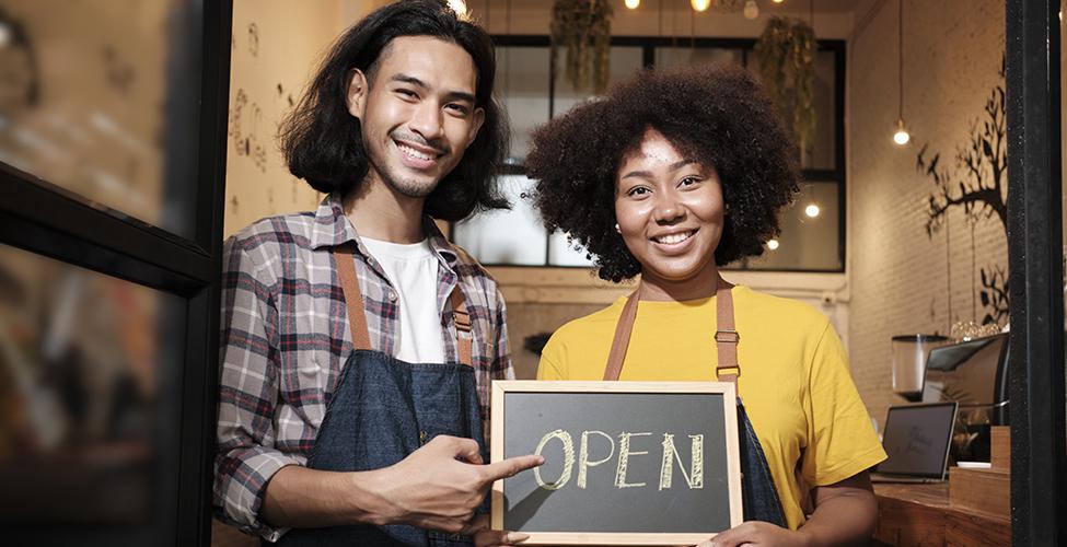 Two young startup barista partners with aprons stand at casual cafe door, letters on board and show open sign