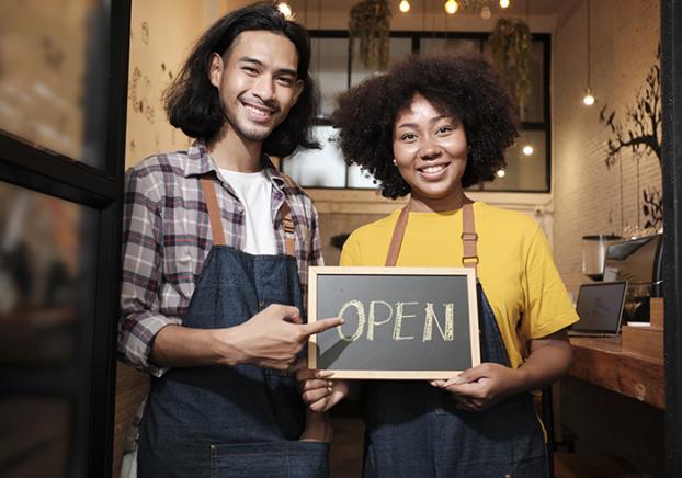 Two young startup barista partners with aprons stand at casual cafe door, letters on board and show open sign